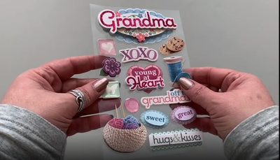 Female hands pick up and show front and back of 3D scrapbook stickers featuring yarn, cookies and words of grandma love.