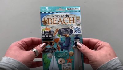Female hands pick up and show front and back of 3D scrapbook stickers featuring photo real lighthouse, sea shells, sailboat and sunglasses.