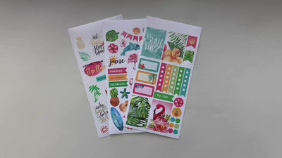 Female hands pick up and show in detail 3 sheets of stickers featuring a tropical June theme.
