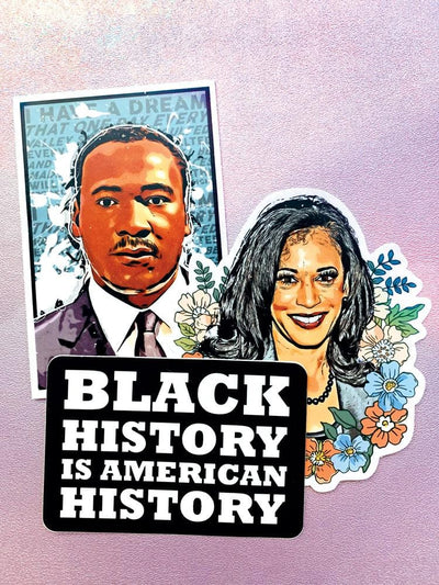 Three laptop stickers are shown on a lavender, metallic background featuring an illustrated Kamala Harris, an illustrated Martin Luther King and "BLACK HISTORY IS AMERICAN HISTORY" in white letters on a black background. 