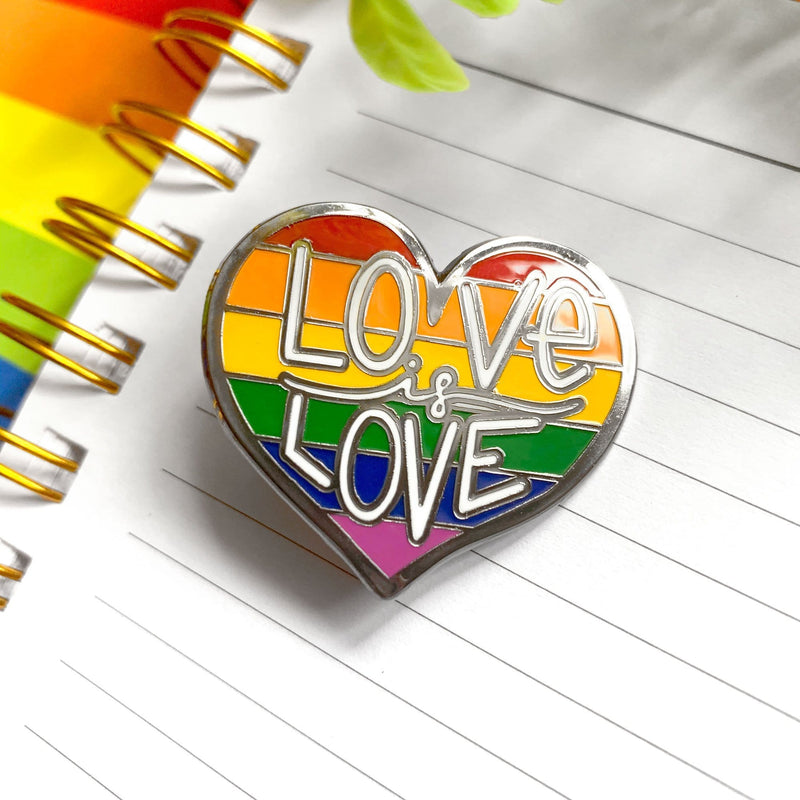 Love is Love enamel pin made of enamel and silver shown on a lined, spiral notebook on a rainbow colored background.