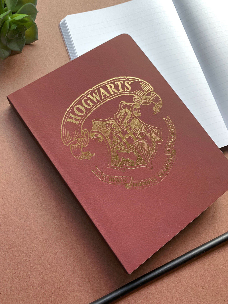 Harry Potter Hogwarts Crest softcover journal notebook shown laying on a desk. Journal cover features a Hogwarts crest printed in gold foil against a dark read background beige background.