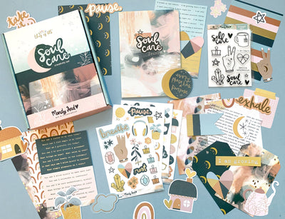 An assortment of craft kit components including papers, stickers, die cuts and envelopes featuring navy, gold and pink patterns and illustrations shown on a blue background.