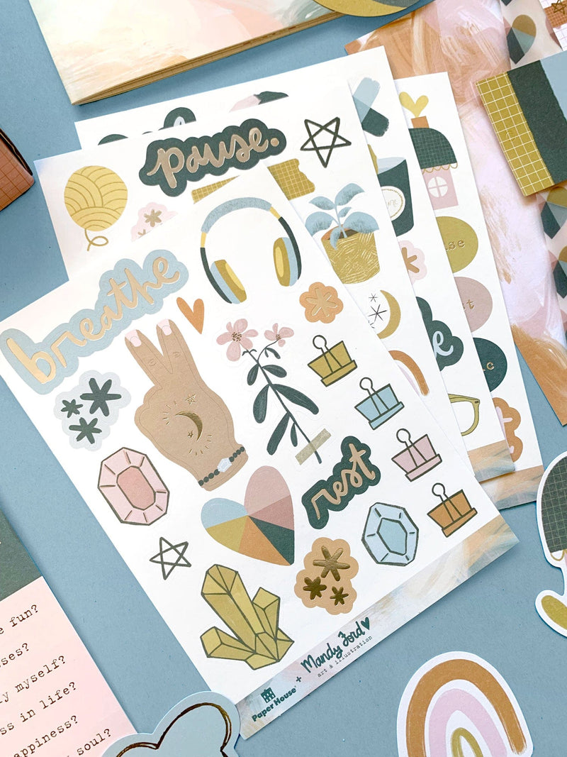 A close up of craft kit components including stickers and die cuts featuring navy, gold and pink patterns and illustrations shown on a blue background.