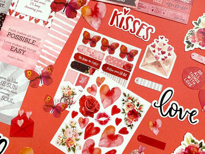 this craft kit closeup image features red, black and pink themed scrapbook papers, stickers, and diecuts featuring hearts, roses and words shown on a red background.