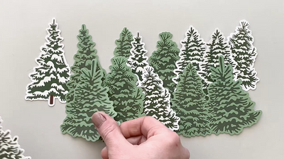 hands display an assortment of die cut, illustrated scrapbook stickers featuring pine trees and pine cones.
