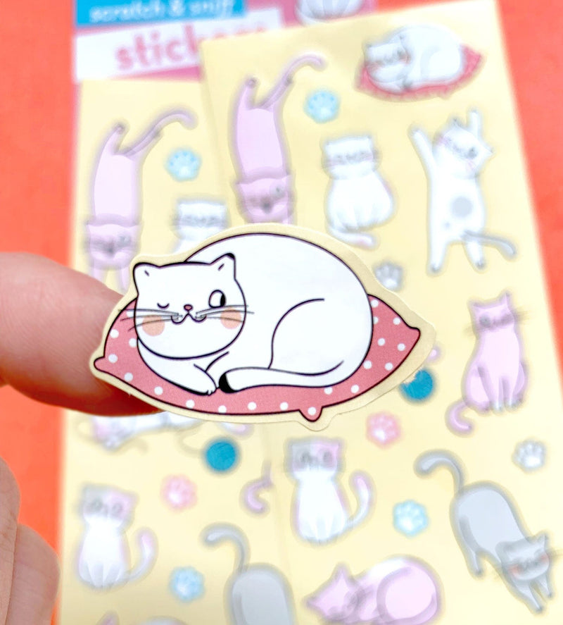 close up of scratch and sniff sticker featuring an illustrated cat on a pillow displayed on finger above a full sheet of stickers in the background.