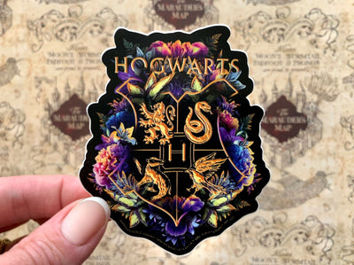 close up of shaped Harry Potter laptop sticker, shown in hand, featuring the Hogwarts crest with floral and black and gold details, shown over a beige pattern of the  marauder's map.