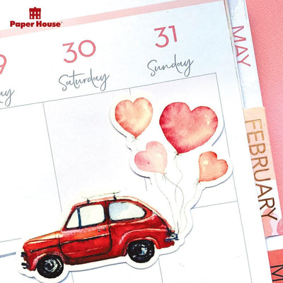 Shaped laptop sticker featuring a red illustrated car towing pink heart-shaped balloons, shown on a white, weekly planner spread featuring the February tabl.