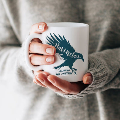 Shaped, vinyl sticker featuring a blue raven with the words Ravenclaw, learning, with and wisdom shown on a white mug held by a person in a grey sweater.