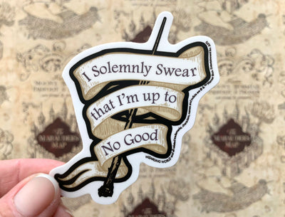 close up of shaped vinyl laptop sticker featuring Harry Potter Solemnly Swear Banner, held in hand over a background of the marauder's map.