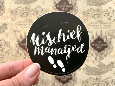 close up of vinyl laptop sticker featuring Harry Potter Mischief Managed on black, circle background, shown in hand over a background of the marauder's map.