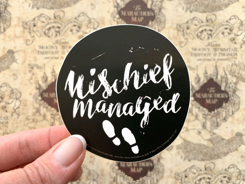 close up of vinyl laptop sticker featuring Harry Potter Mischief Managed on black, circle background, shown in hand over a background of the marauder&