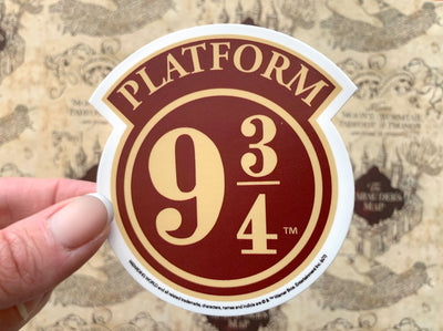 close up of vinyl laptop sticker featuring Harry Potter Platform 9 3/4 sign, held in hand over a marauder's map background.