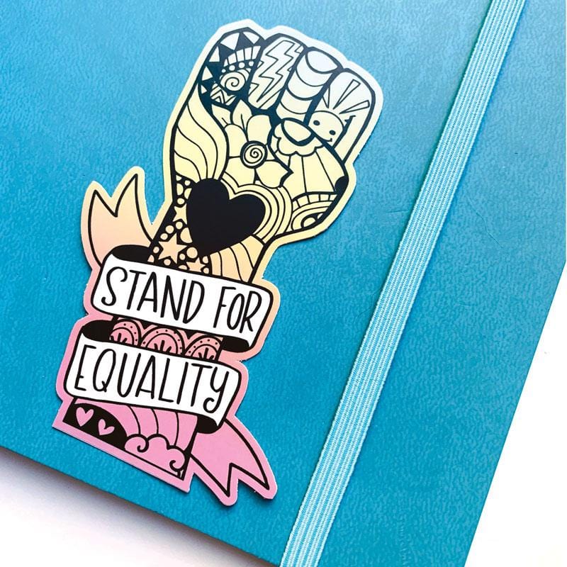 Shaped laptop sticker featuring a colorful, illustrated power fist with a "Stand for Equality" banner wrapped around it, shown on a blue notebook with elastic band.