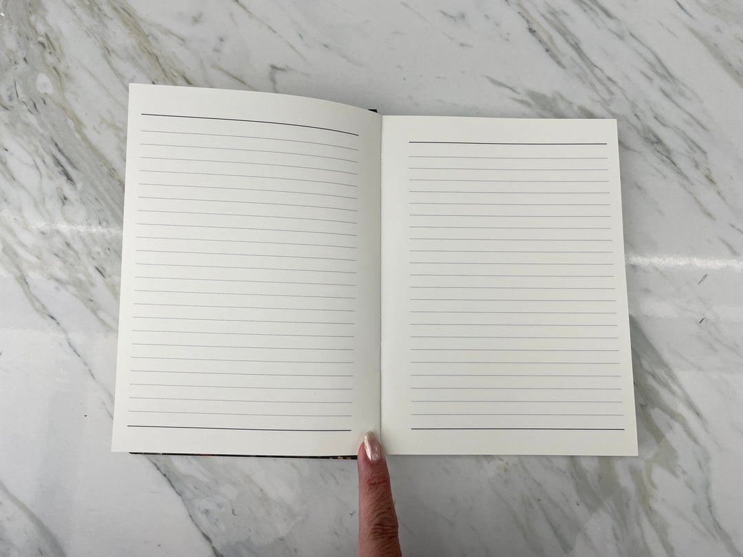 JL-0082E - Skull &amp; Flowers - Lined Journal - Open lined notebook with blank pages displayed on marble surface, finger pointing to center binding