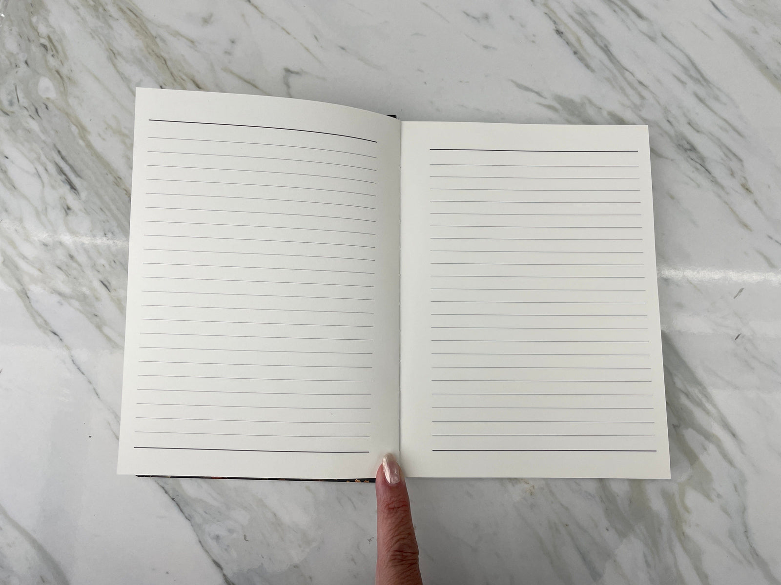 JL-0082E - Skull & Flowers - Lined Journal - Open lined notebook with blank pages displayed on marble surface, finger pointing to center binding