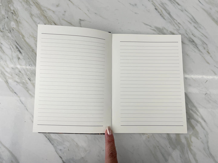 JL-0082E - Skull &amp; Flowers - Lined Journal - Open lined notebook with blank pages displayed on marble surface, finger pointing to center binding