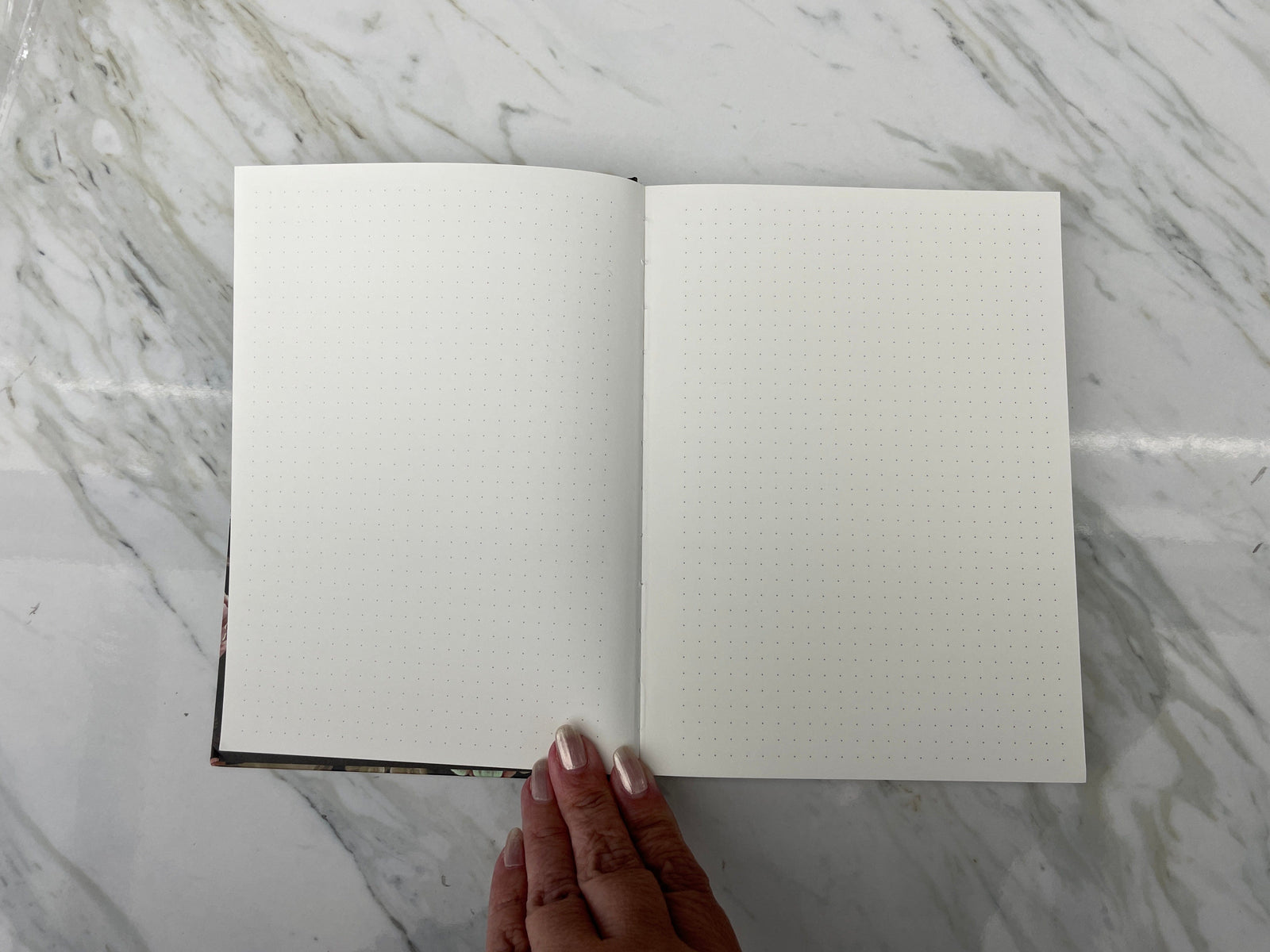 JL-0086E - Skulls & Flowers Pattern Dot Grid Journal - Open dot grid journal with cream pages and dark patterned spine, held by hand against marble surface