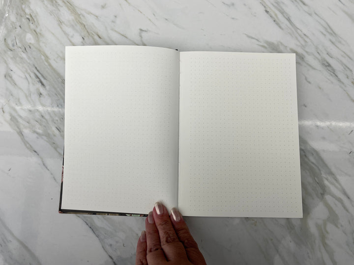 JL-0086E - Skulls &amp; Flowers Pattern Dot Grid Journal - Open dot grid journal with cream pages and dark patterned spine, held by hand against marble surface