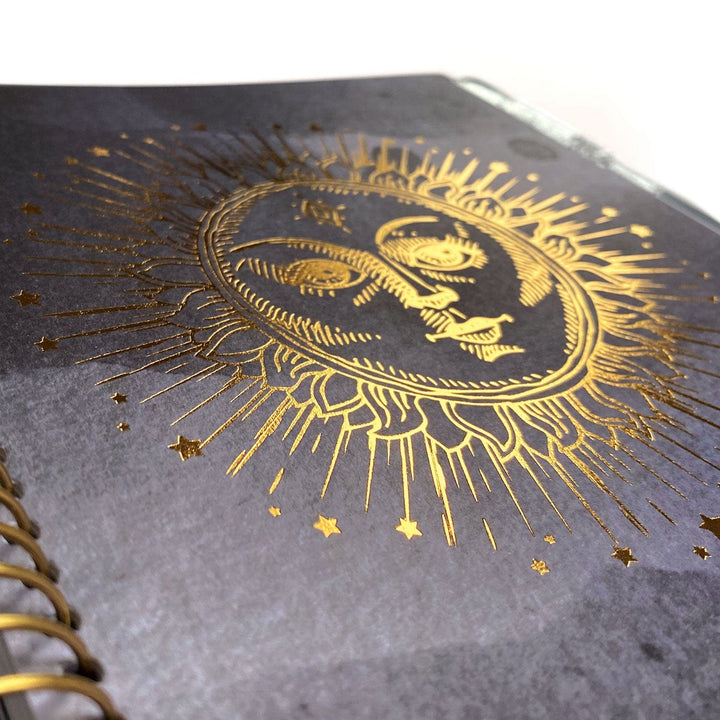 PL-2016E - Weekly Planner - Undated Celestial - Gray spiral-bound planner with gold foil celestial sun face design and radiating rays surrounded by stars