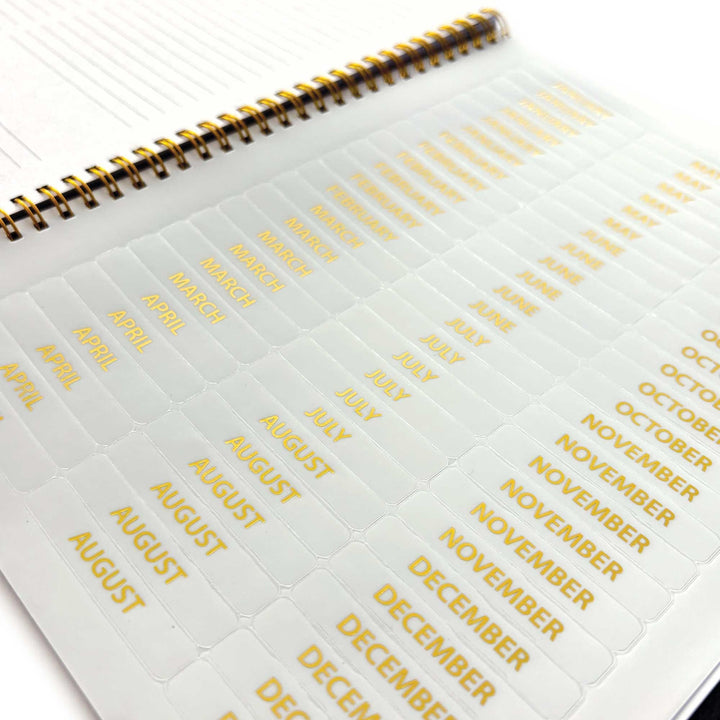 PL-2036E - Books &amp; Flowers 12 Month Undated Planner - Gold spiral-bound planner with white pages showing month tabs labeled April through December in gold text
