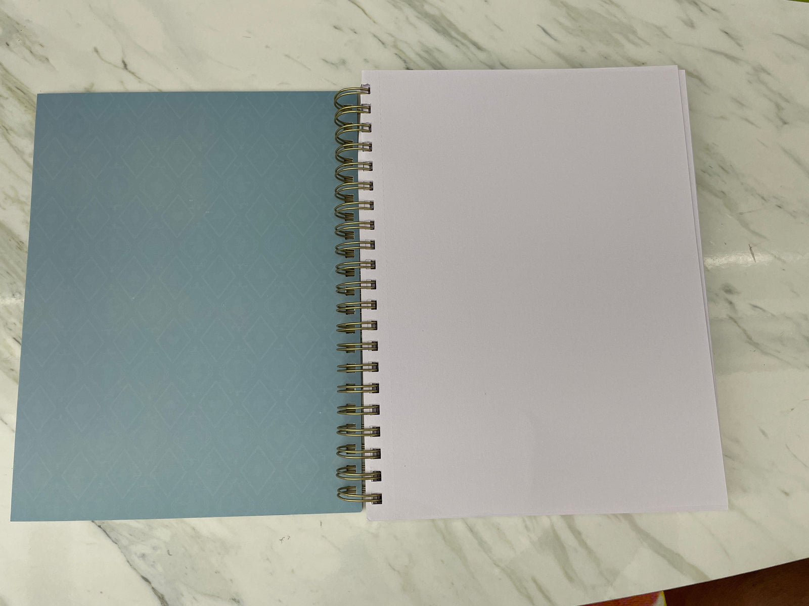 SKB-0003E - Book Shelves Sketchbook - Open spiral-bound notebook with light blue patterned cover and blank white pages on marble surface
