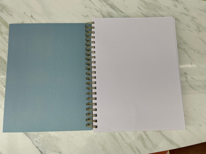 SKB-0003E - Book Shelves Sketchbook - Open spiral-bound notebook with light blue patterned cover and blank white pages on marble surface