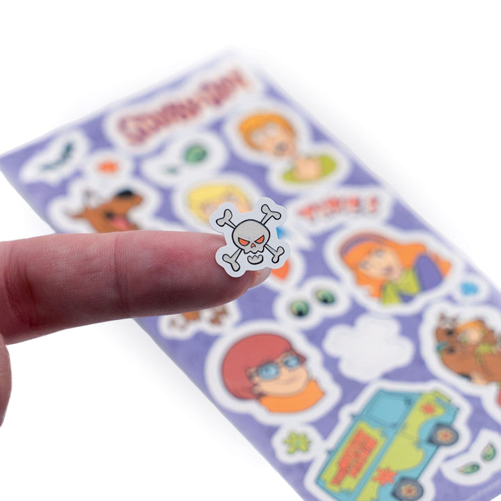 STGD-7007E - Glow In the Dark Stickers - Scooby Doo - Glow-in-the-dark skull and crossbones sticker being held above a purple sheet of colorful character stickers