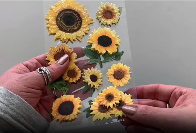 Female hands pick up and show front and back of photo real 3D decorative stickers featuring sunflowers.