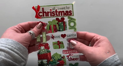 Female hands pick up and show front and back of photo real 3D scrapbook stickers featuring red and green Christmas presents.