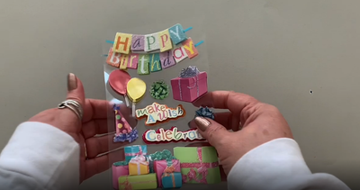 Female hands pick up and show front and back of 3D scrapbook stickers featuring birthday presents, banners and balloons.