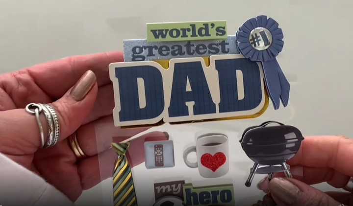 Female hands pick up and show front and back of 3D scrapbook stickers featuring sports balls, a tie, a grill and dad words.