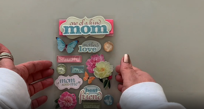Female hands pick up and show front and back of 3D scrapbook stickers featuring pastel flowers and words of love for mom.