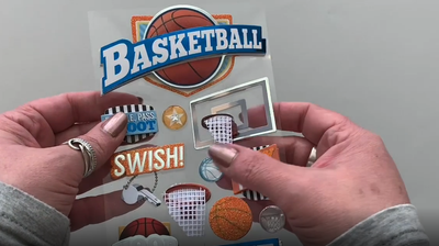 Female hands pick up and show front and back of 3D scrapbook stickers featuring basketballs and hoops with orange and blue foil details. 