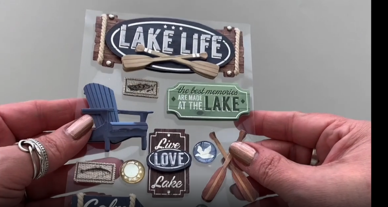 Female hands pick up and show front and back of 3D scrapbook stickers featuring a lake life theme with  a canoe and adirondack chair.