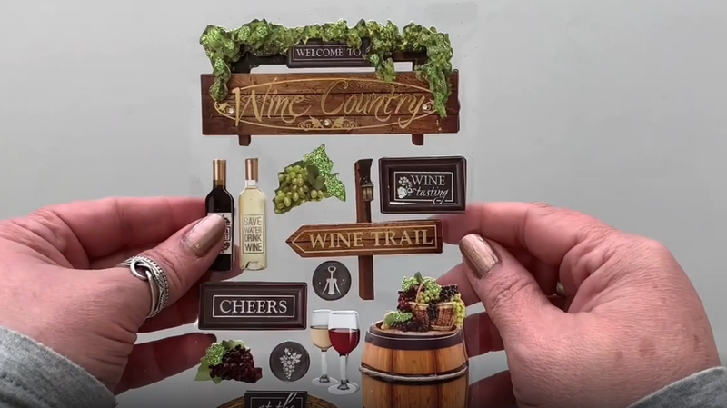 Female hands pick up and show front and back of 3D scrapbook stickers featuring photo real grapes, wine bottles and wine glasses.