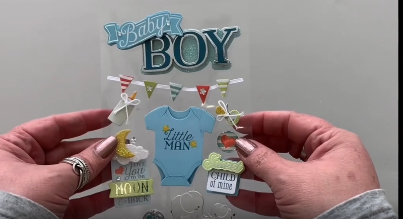 Female hands pick up and show front and back of 3D scrapbook stickers featuring a baby onesie, a bottle and silver elephant.