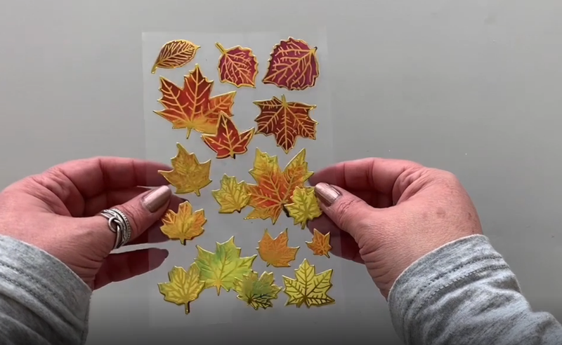 Female hands pick up and show front and back of 3D scrapbook stickers featuring fall leaves with gold foil accents.