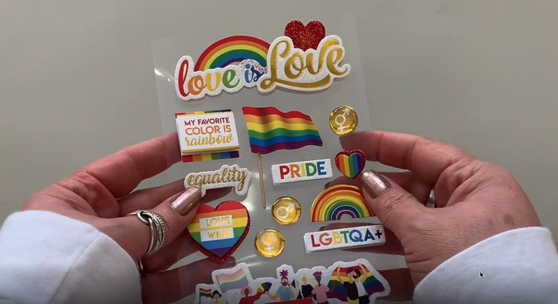 Female hands pick up and show front and back of 3D scrapbook stickers featuring rainbow illustrations and words of pride and "love is love"