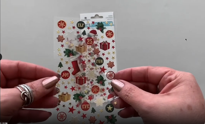 Female hands picking up and showing front and back of a sheet of mini stickers featuring gold, red and green snowflakes, stars and christmas presents.