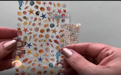 Female hands showing a sheet of mini stickers featuring colorful seashells and placing them on surface with package and another sheet of mini stickers.