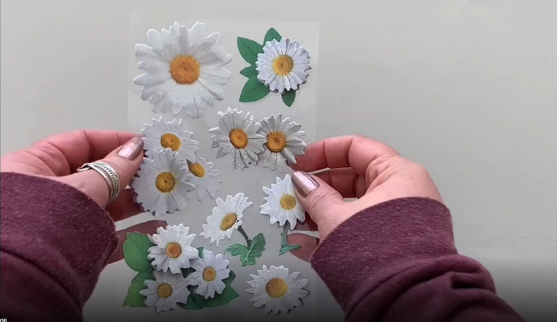 Female hands pick up and show in detail 3D scrapbook stickers featuring photo-real yellow and white oxeye daisies.
