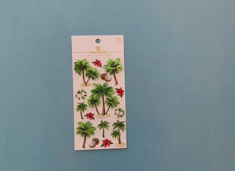 female hands display scrapbook stickers featuring green palm trees and tropical flowers with green foil and shows close up of 2 individual stickers.