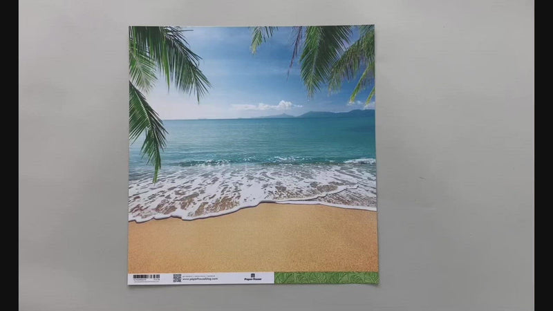 Female hands pick up and show scrapbook paper featuring a tropical beach photo on one side and a green, illustrated leaf pattern on the other side.