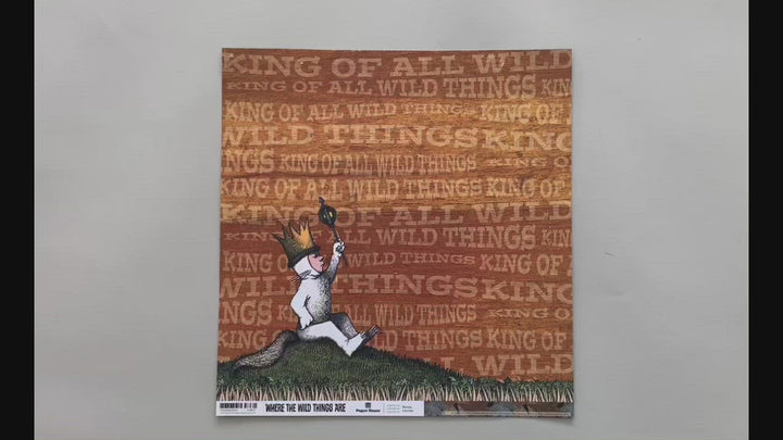 Female hands pick up scrapbook paper featuring Max from Where the Wild Things Are sitting on a hill on one side and a pattern of illustrated leaves on the other side.