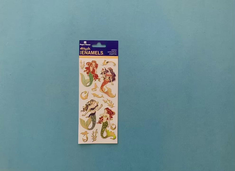 female hands display sheet of foil stickers featuring colorful, illustrated mermaids with gold details and shows detail of a single sticker.