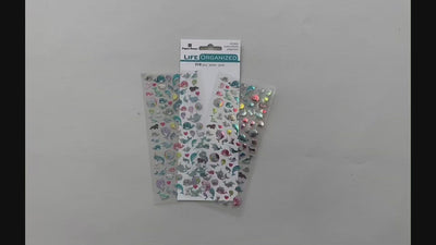 Female hands showing front and back of sheet of mini stickers featuring pastel colored narwhals. And placing them on surface with package and another sheet of stickers. 