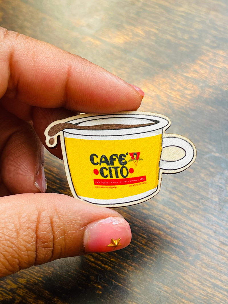 enamel pin image featuring yellow and gold cafecito coffee mug held by hands, shown on wooden surface.