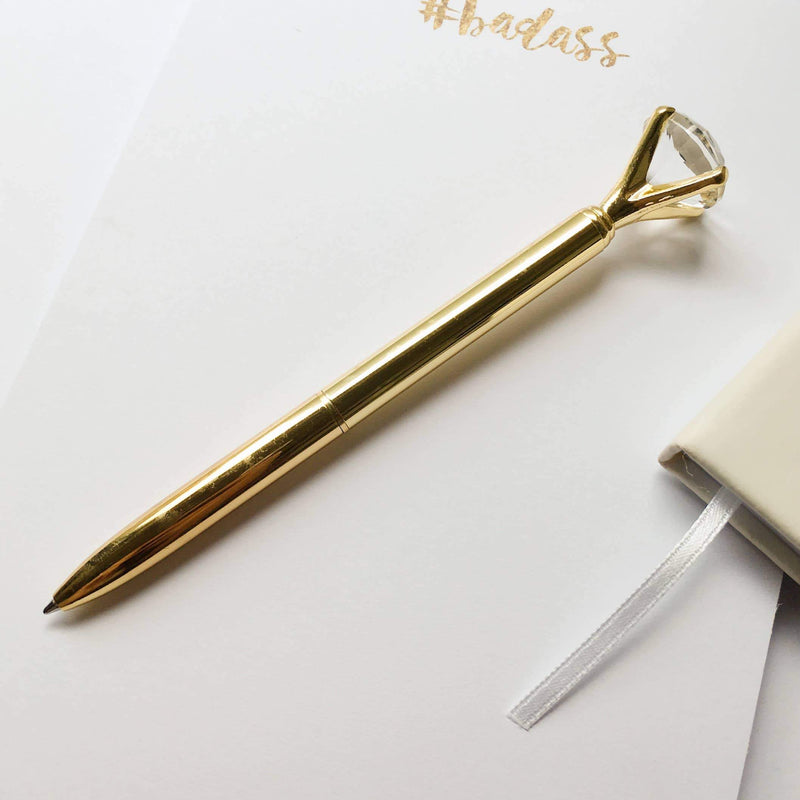 gold pen featuring a diamond rhinestone at top, shown on white notepaper with white journal and ribbon.